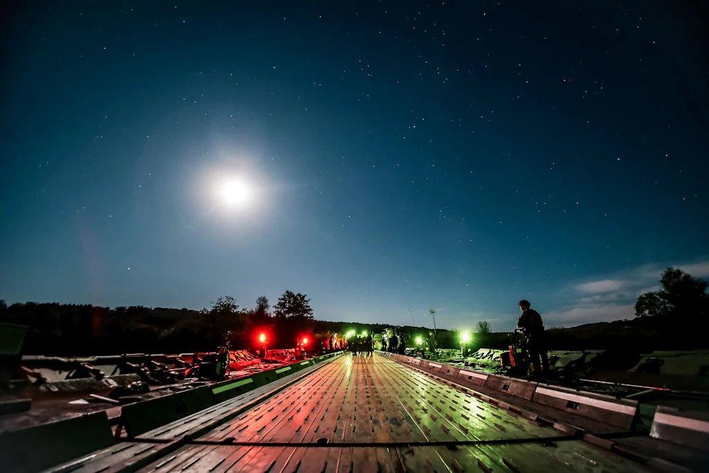 Blick entlang einer beleuchteten Schwimmbrücke unter dem Mond und einem sternenklaren Himmel; mehrere Armeeangehörige stehen auf der Brücke.
