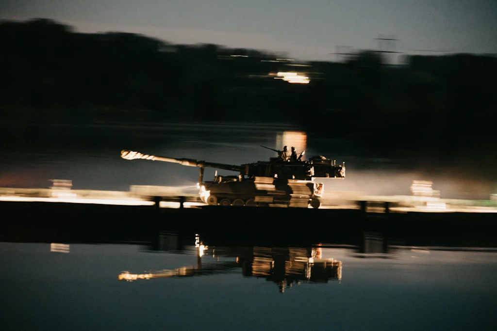 Eine Panzerhaubitze M109 fährt bei Nacht über eine beleuchtete Schwimmbrücke; zwei Besatzungsmitglieder sind auf dem Fahrzeug sichtbar.
