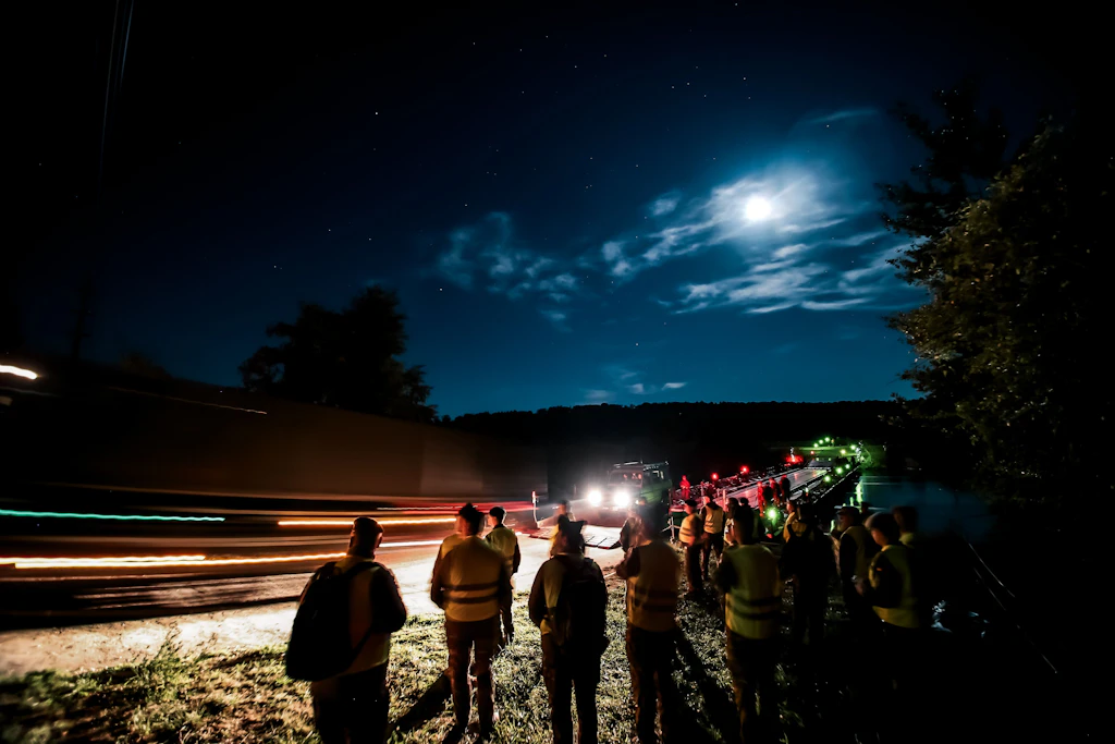 Bei Mondlicht überquert ein Militärfahrzeug eine beleuchtete Schwimmbrücke. Am Ufer beobachten mehrere Personen in Warnwesten den nächtlichen Übergang.