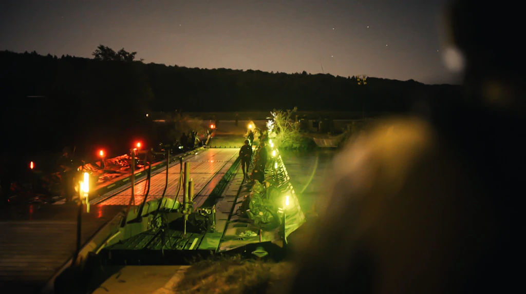 Eine beleuchtete Schwimmbrücke bei Nacht; mehrere Armeeangehörige stehen auf und neben der Brücke.
