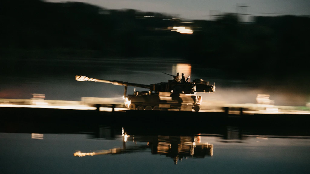 Un obusier blindé M109 traverse de nuit un pont flottant éclairé; deux membres de l’équipage sont visibles sur le véhicule.