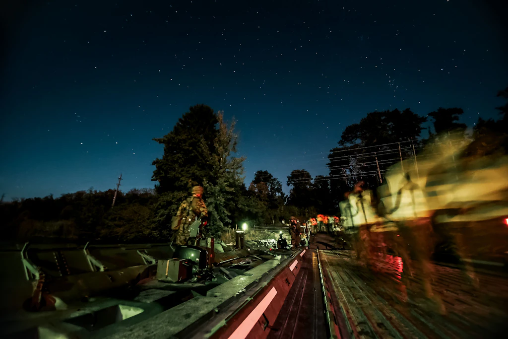 Un véhicule militaire traverse de nuit un pont flottant éclairé, tandis que des militaires accompagnent son passage.