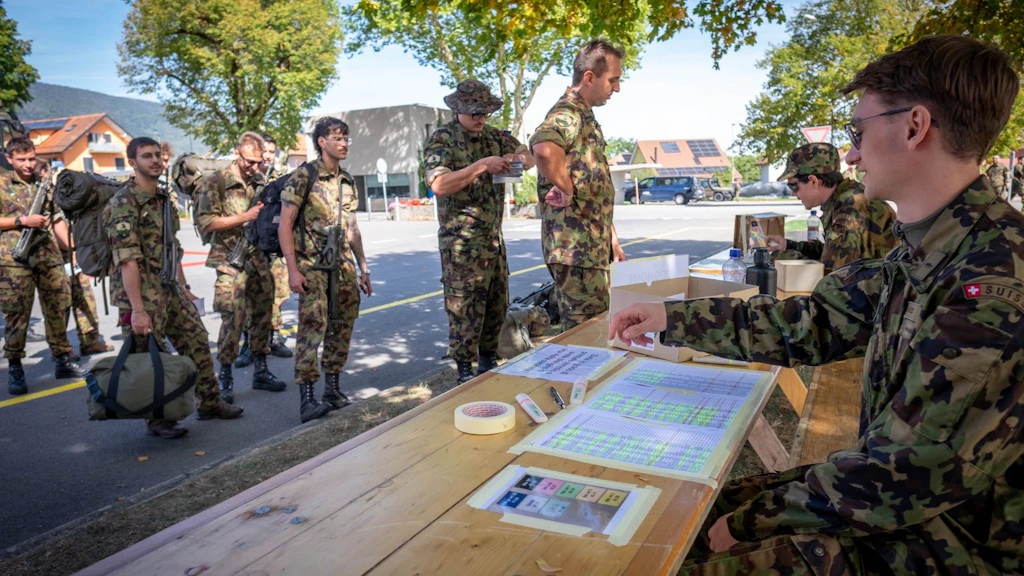 Alcuni militari con il proprio bagaglio attendono davanti a un tavolo di controllo all’aperto, dove altri militari verificano i documenti.