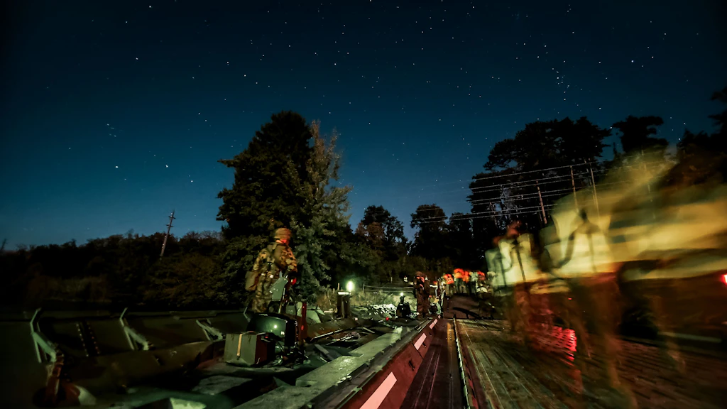 Un veicolo militare attraversa di notte un ponte galleggiante illuminato, mentre alcuni militari ne accompagnano il passaggio.