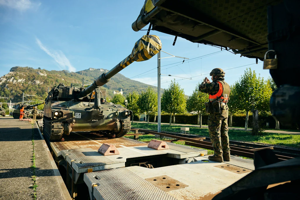 Un militare guida un obice blindato M109 su un carro ferroviario; sullo sfondo attendono altri veicoli cingolati.