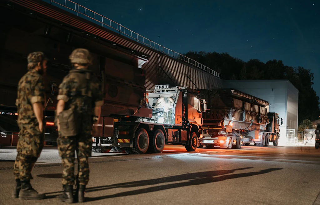 Due militari si trovano di notte davanti ad autocarri militari pesanti illuminati e carichi di elementi del ponte.