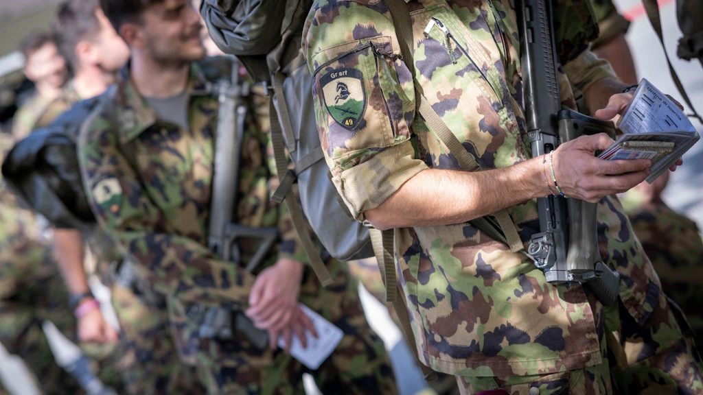 Primo piano di un militare con fucile d’assalto e documenti personali; sulla manica è visibile il distintivo del gruppo d’artiglieria 49.